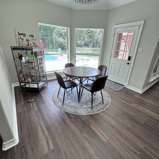 Modern breakfast nook with dark wood flooring, a round dining table with leather chairs, a statement chandelier, and large windows offering a view of the backyard pool. Home renovation and flooring services available in Kingwood, Houston, and Katy.